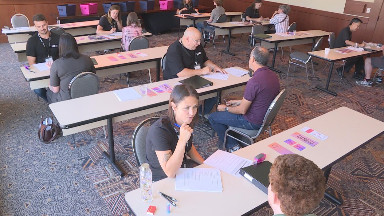 Vue sur quelques tables dans une grande salle. À chacune des tables, il y a deux personnes qui discutent ensemble. Il y a des papiers sur les tables.