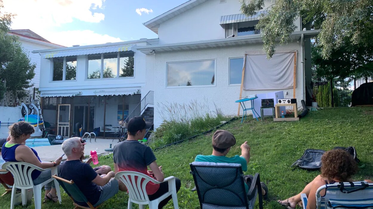 Cinq personnes sont assis sur des chaises et regardent un écran installé à l'extérieur dans la cour arrière d'une maison.