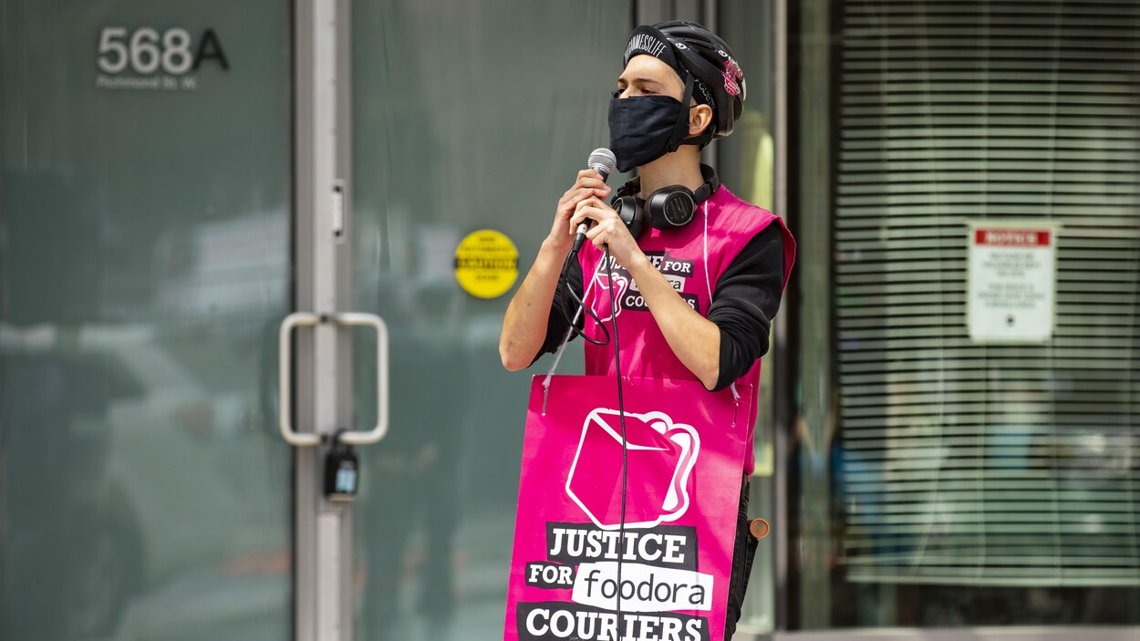 Un manifestant avec un masque au visage tient un micro dans ses mains et porte une affiche sur laquelle il est inscrit « Justice for Foodora Couriers ».