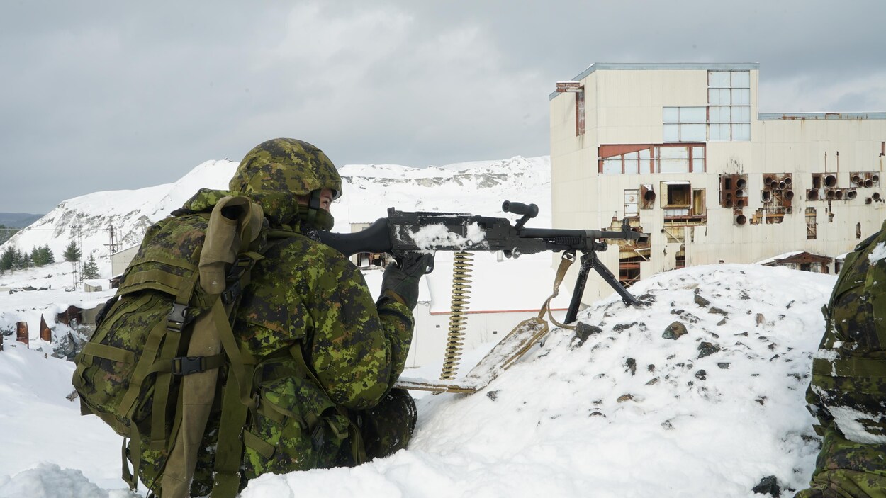 Un militaire tient un fusil mitraillette avec une ceinture de balles avec en arrière-plan une mine abandonnée.