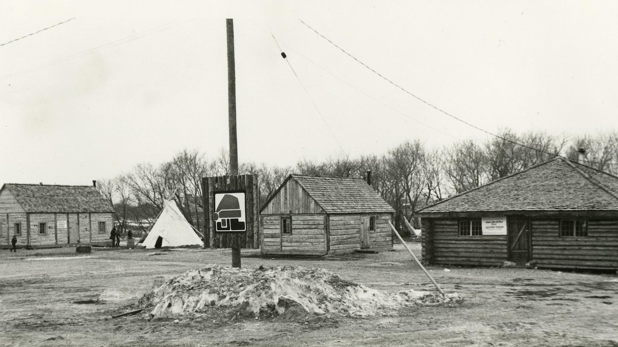 4 cabanes de bois rond dans la neige. Entre la deuxième et la troisième se dresse un tipi.