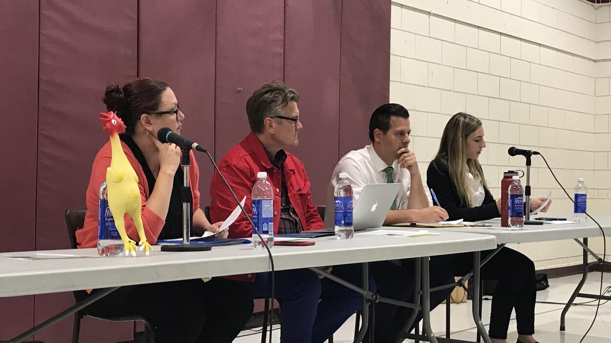 Quatre personnes assises devant une grande table, avec des micros, dans une salle communautaire.