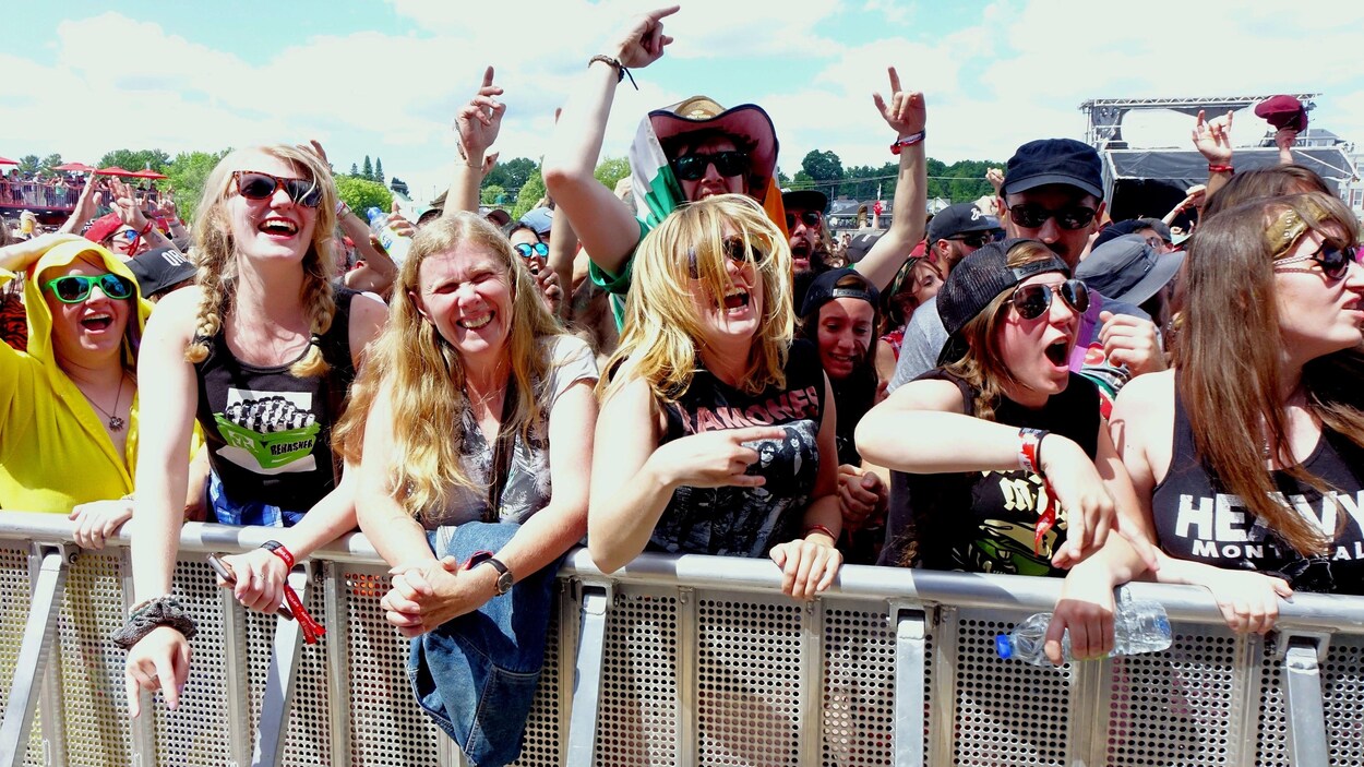Des femmes souriantes et enthousiastes sont alignées le long d'une clôture lors d'un spectacle du Rockfest en plein soleil.