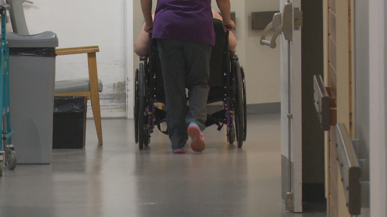 Une préposée aide un résident à se déplacer en fauteuil roulant dans un couloir du foyer de soins.