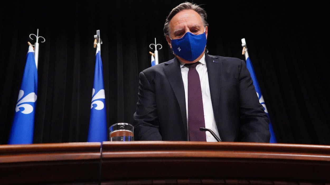 François Legault, debout derrière un bureau et devant des drapeaux du Québec, portant un masque.