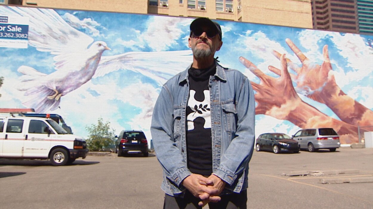 Doug Driediger devant une fresque où deux mains sont tendues vers une colombe en vol.