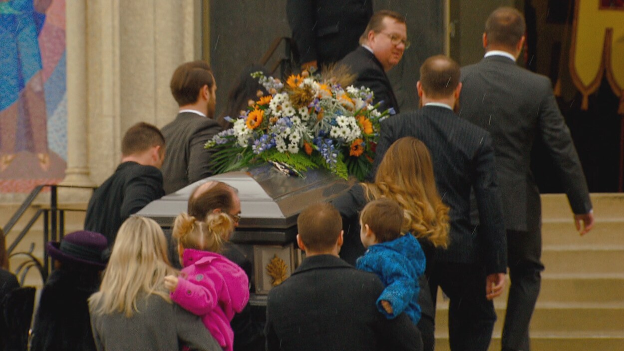 Des hommes transportent un cercueil décoré d'une gerbe de fleurs sur les marches de la cathédrale. De jeunes parents avec des enfants dans les bras suivent le cortège.