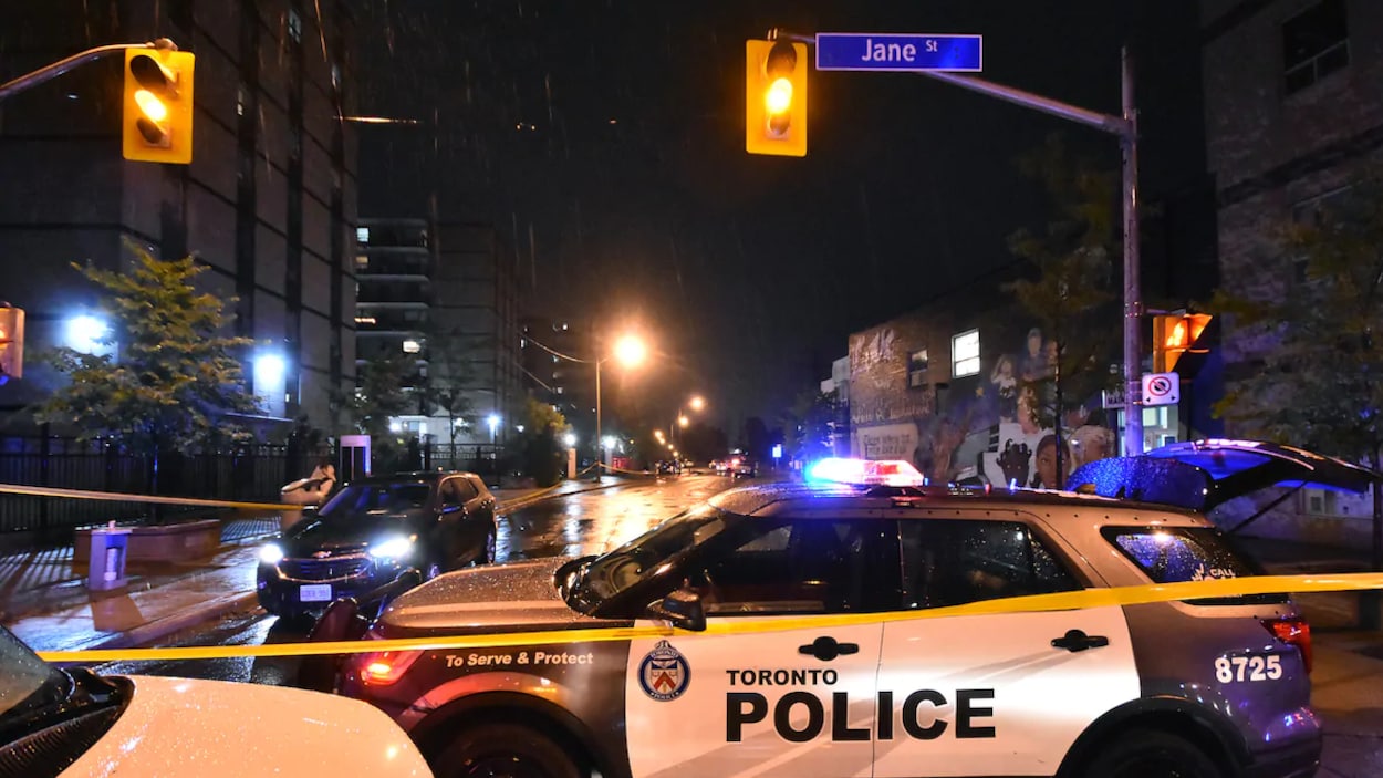 Une scène de crime, la nuit, entourée du ruban jaune de police. Plusieurs véhicules d'urgence sont sur place.