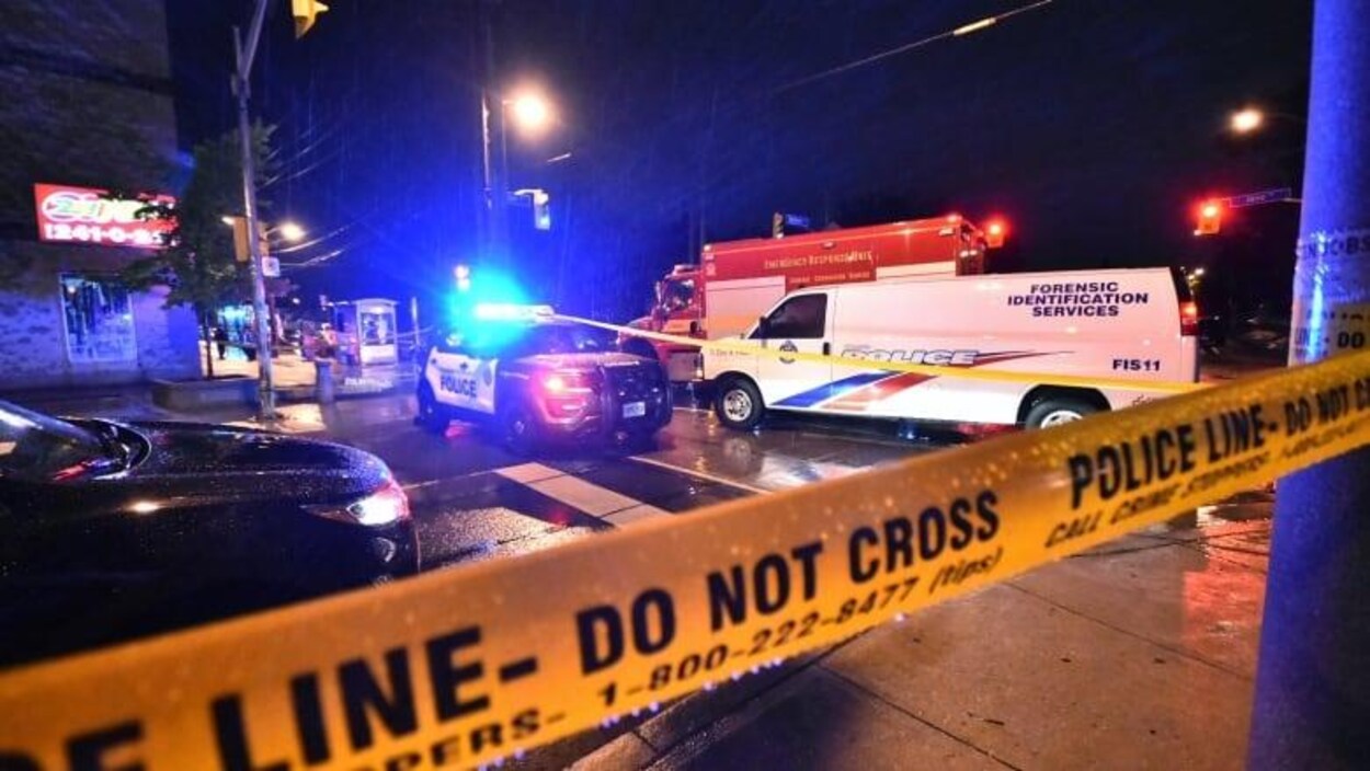 Une scène de crime, la nuit, entourée du ruban jaune de police. Plusieurs véhicules d'urgence sont sur place.