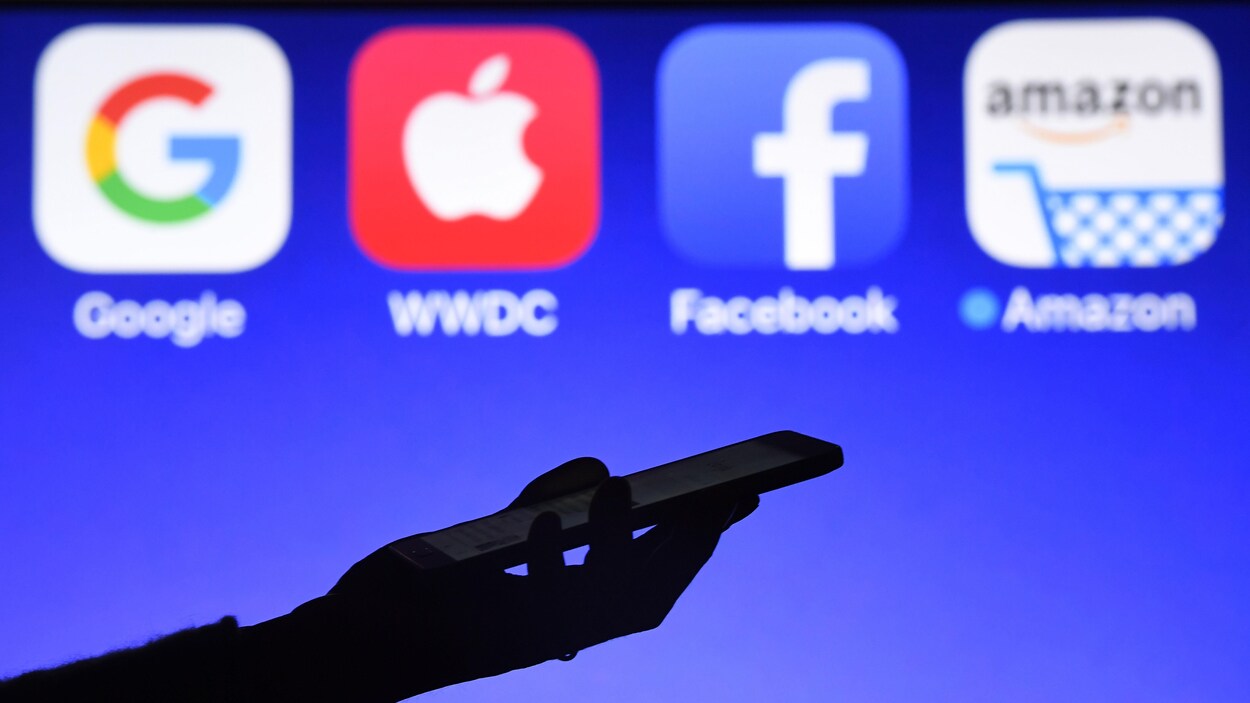 Une personne tient un téléphone devant les logos de Google, Apple, Facebook et Amazon.