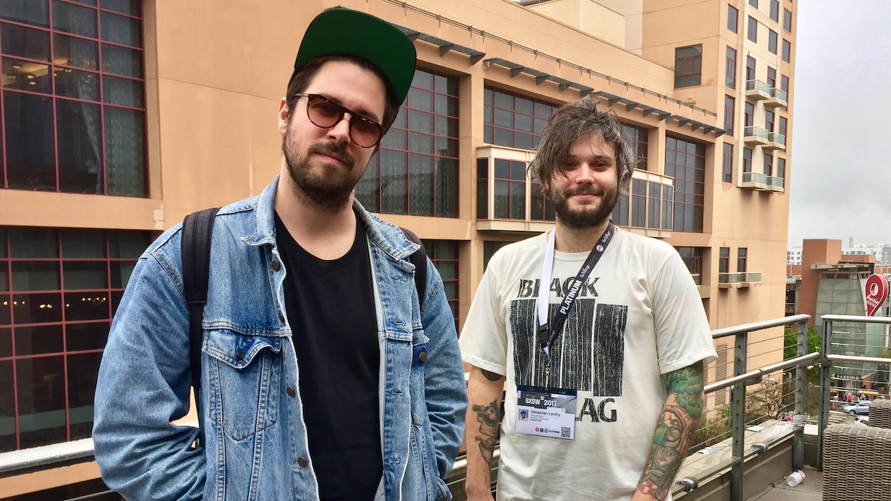 Deux jeunes hommes barbus sourient en regardant dans l'objectif de la caméra.