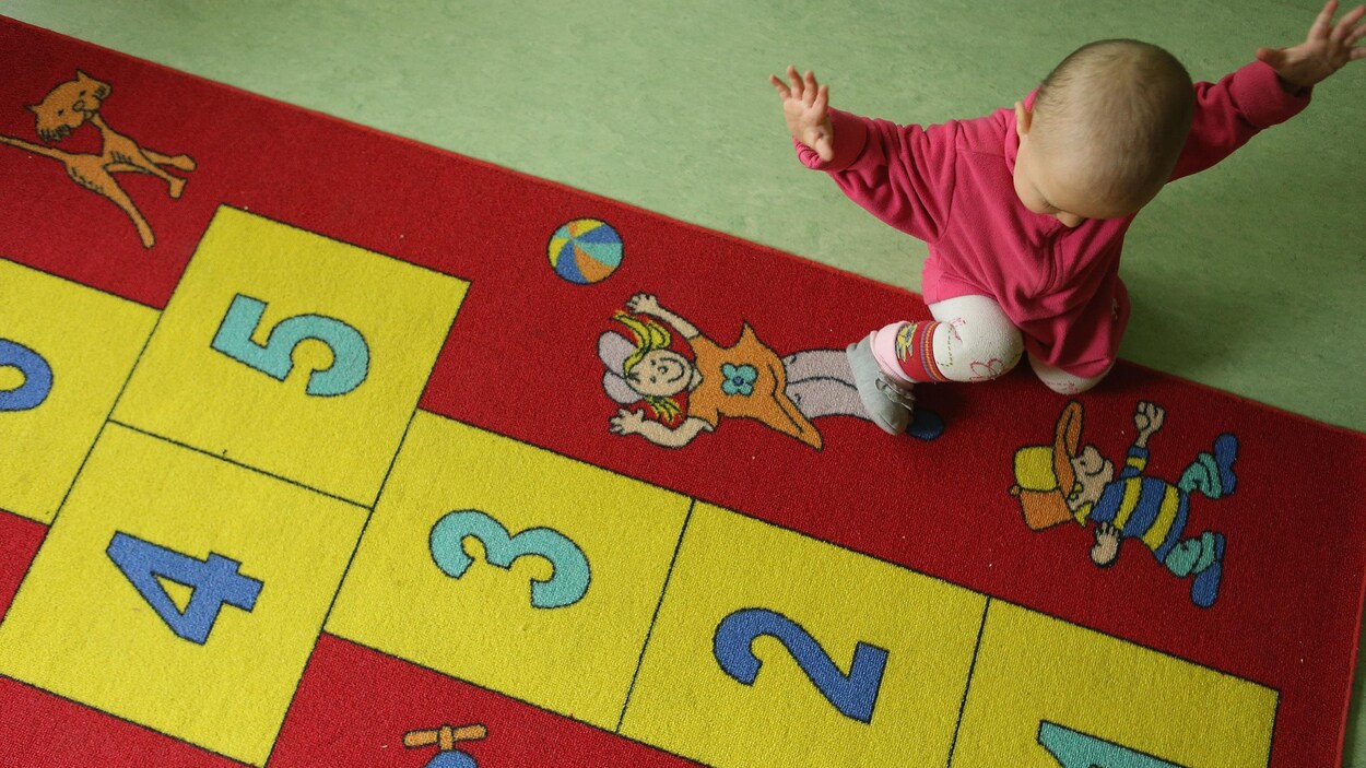 Une enfant de 11 mois, habillée avec une veste rose, s'amuse sur un tapis rouge montrant des personnages qui entoure un jeu de marelle numéroté.