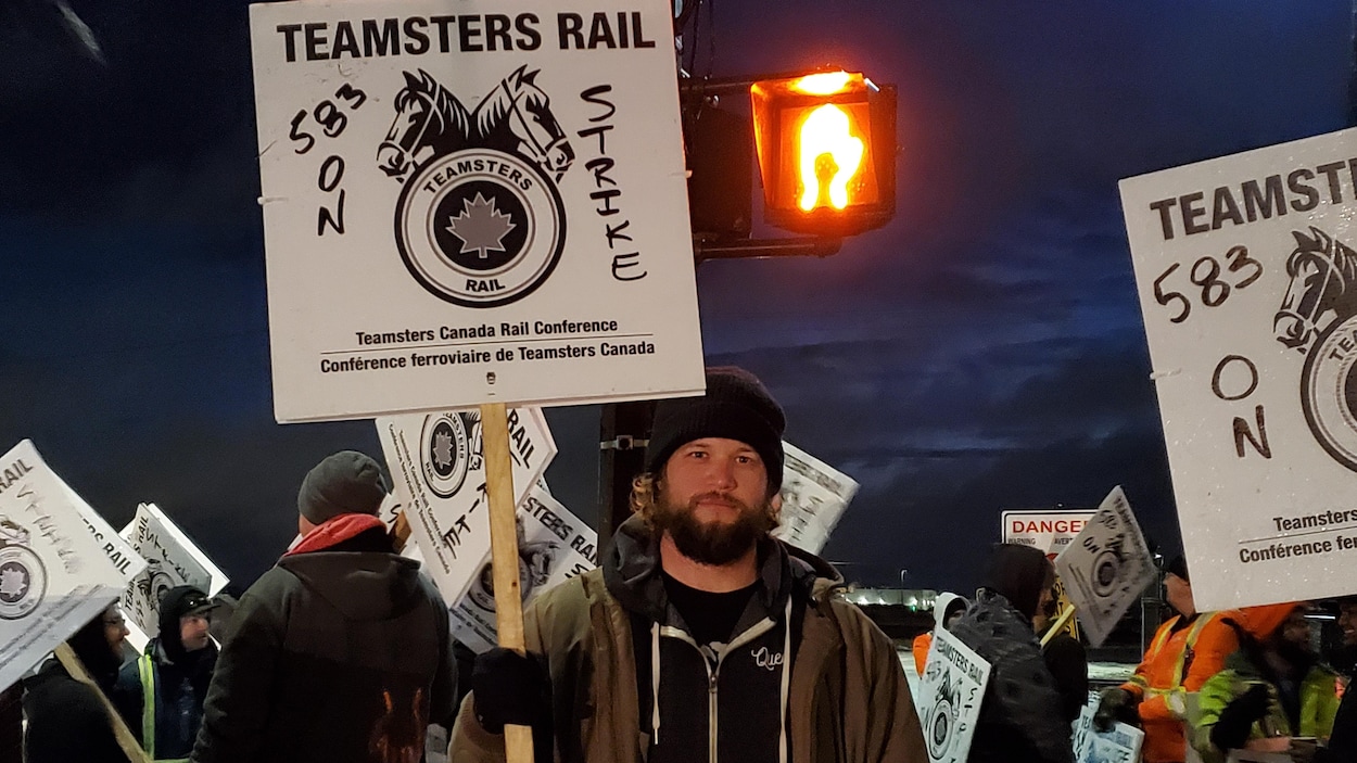 Un homme portant une affiche de son syndicat regarde la photographe. Il est entouré d'autres travailleurs portant aussi des affiches, et le soleil n'est pas encore levé.