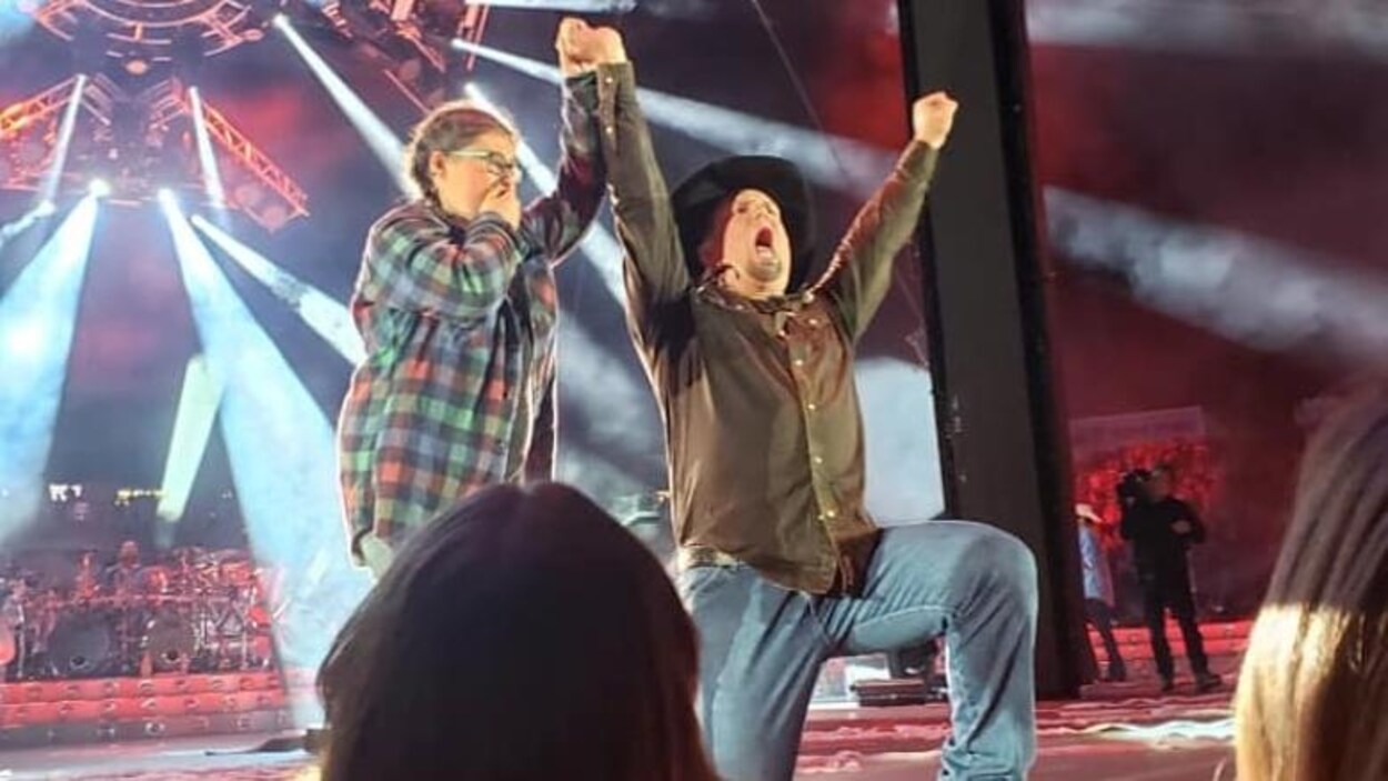 Garth Brooks agenouillé à côté de Camille Baragar sur la scène lève le bras de la jeune fille haut dans les airs.