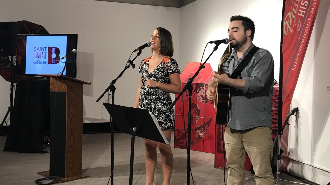 Une femme et un homme chantent au micro dans la galerie d'un musée. Au fond, un écran montre le logo de « Saint-Boniface célèbre ».