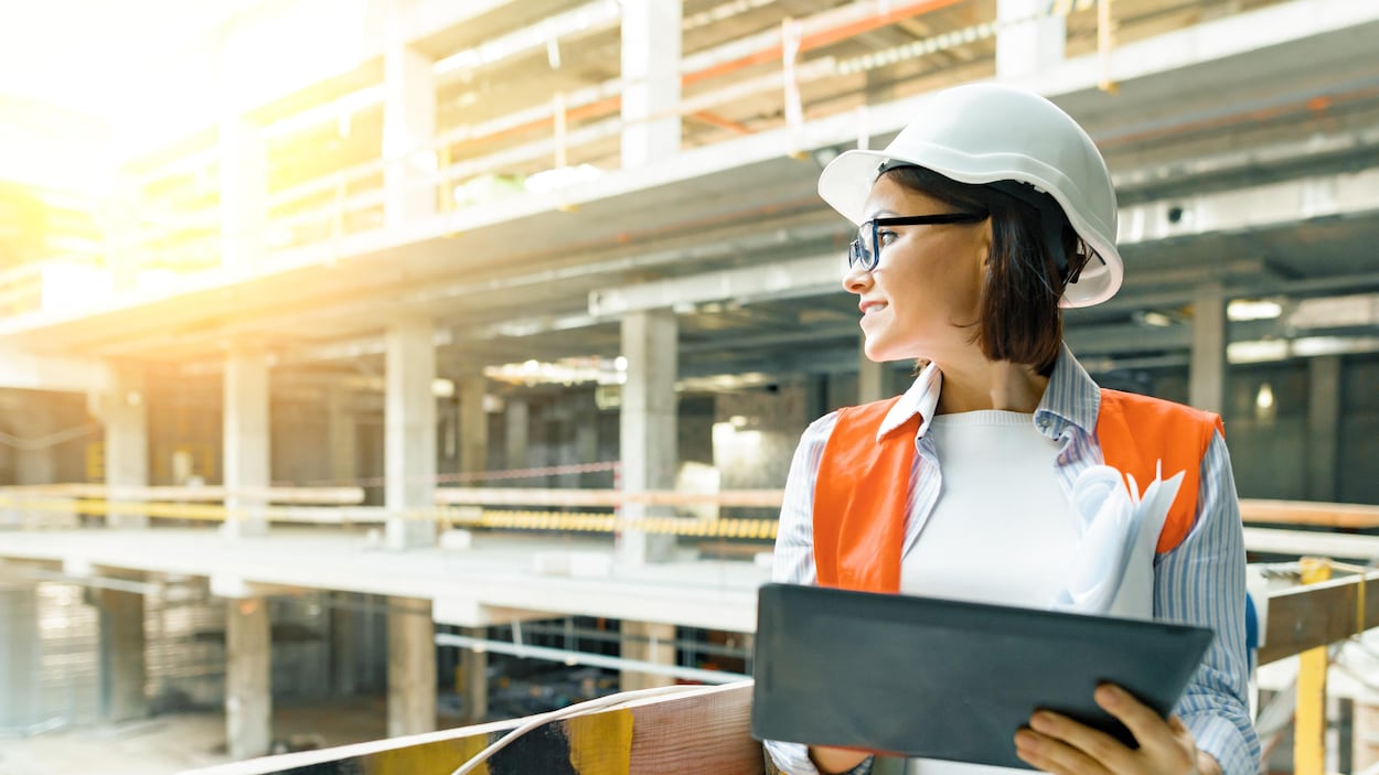 Une femme portant un casque protecteur et un gilet voyant se trouve sur un site en construction. Elle tient également une tablette dans ses mains et porte des lunettes.