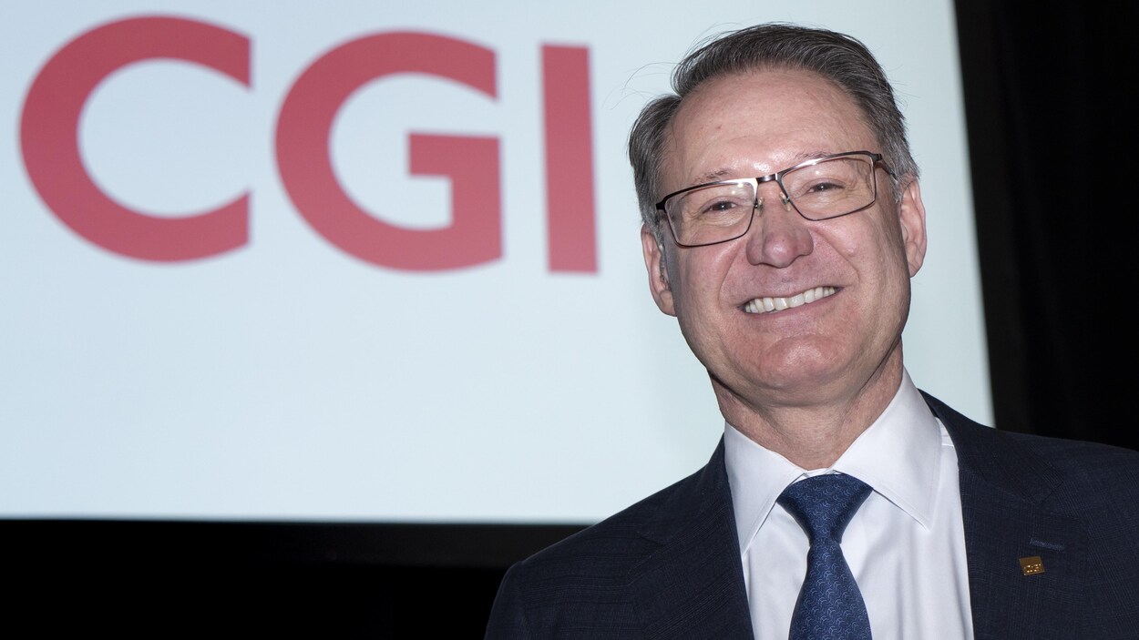 Un homme en complet et portant des lunettes sourit, debout, devant le logo géant CGI projeté sur un écran.