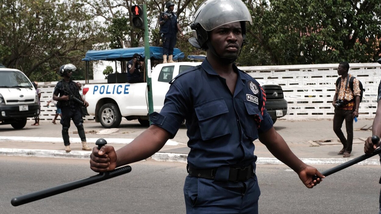 Un policier en uniforme et casqué tient une matraque dans sa main droite.