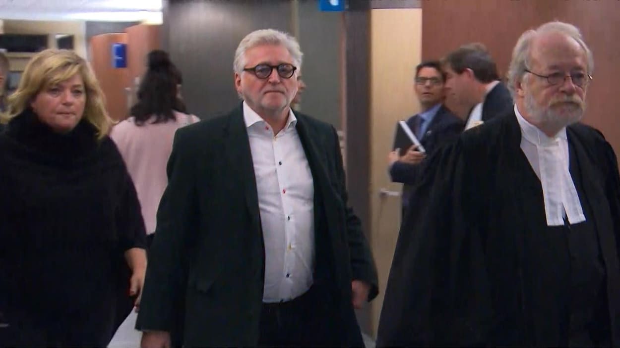 M. Rozon porte des lunettes, un veston et une chemise. Devancé par son avocat, il regarde la caméra, impassible.