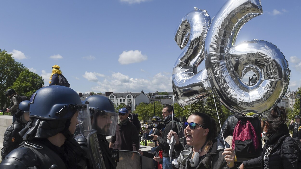 Une femme tenant des ballons en forme du nombre vingt-six s'adresse à des policiers en tenue antiémeute sur une place publique de Nantes.