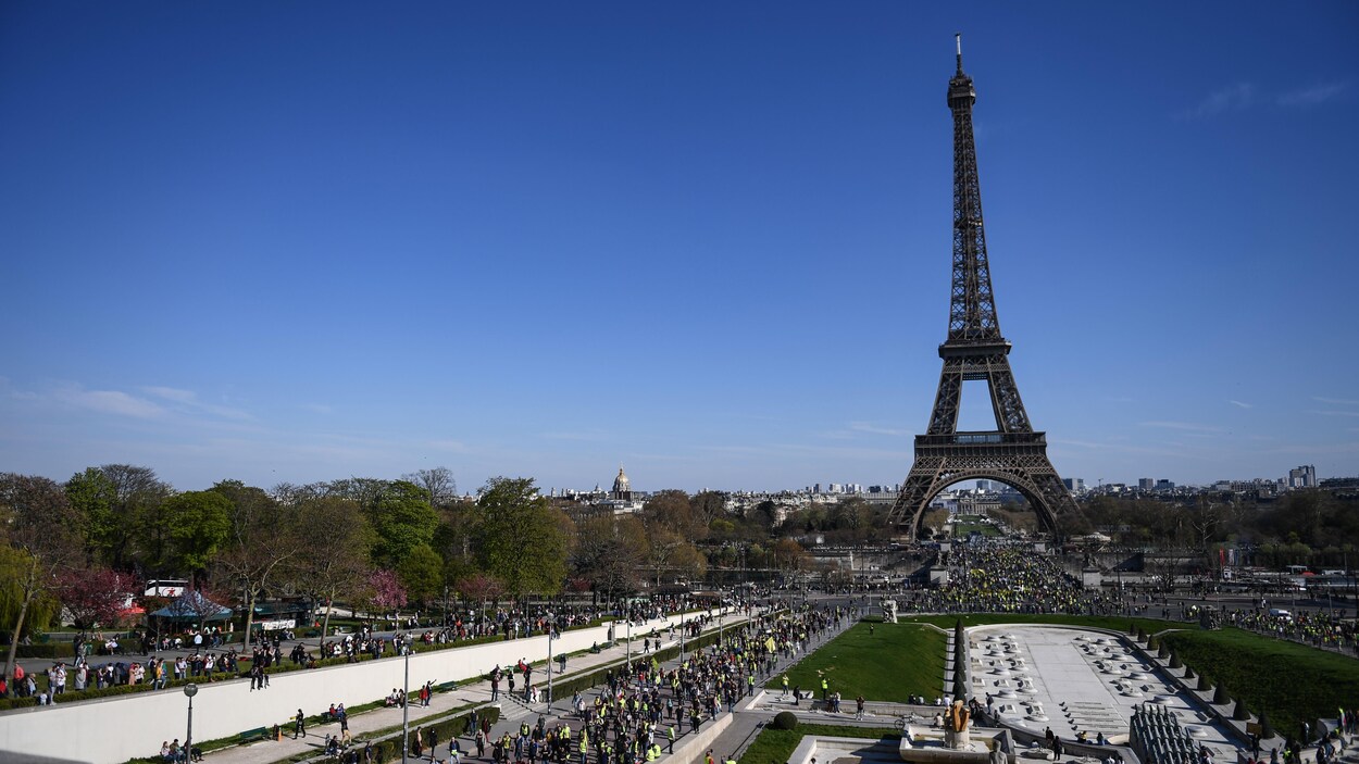 Plusieurs centaines de personnes marchent en direction et sous la tour Eiffel à Paris sous un ciel bleu dégagé.