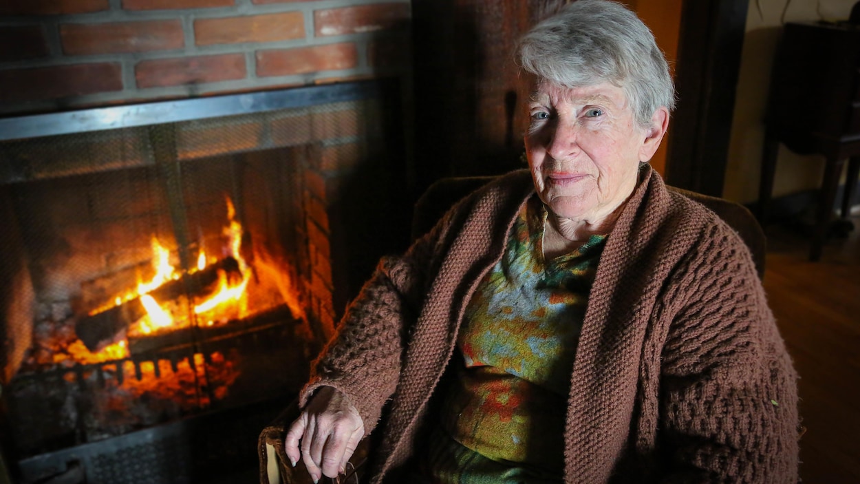 Une dame âgée est assise dans un fauteuil près d'un feu de cheminée.