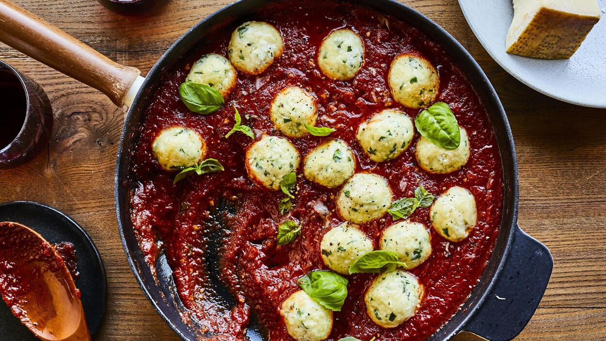 Une poêle remplie de gnudis, de sauce tomate et de basilic repose sur une table à manger, aux côtés d’une spatule tachée de sauce et d’une assiette avec du parmesan.