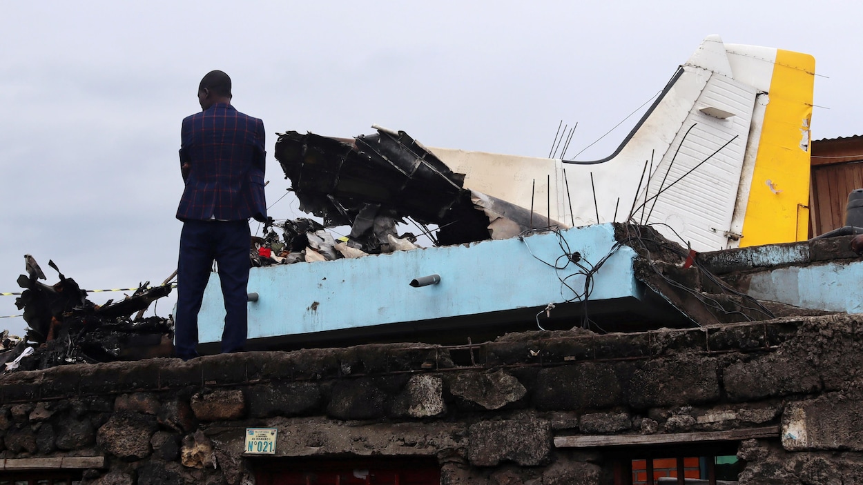 Un homme debout sur le toit endommagé d’une maison à côté de la queue d’un avion.