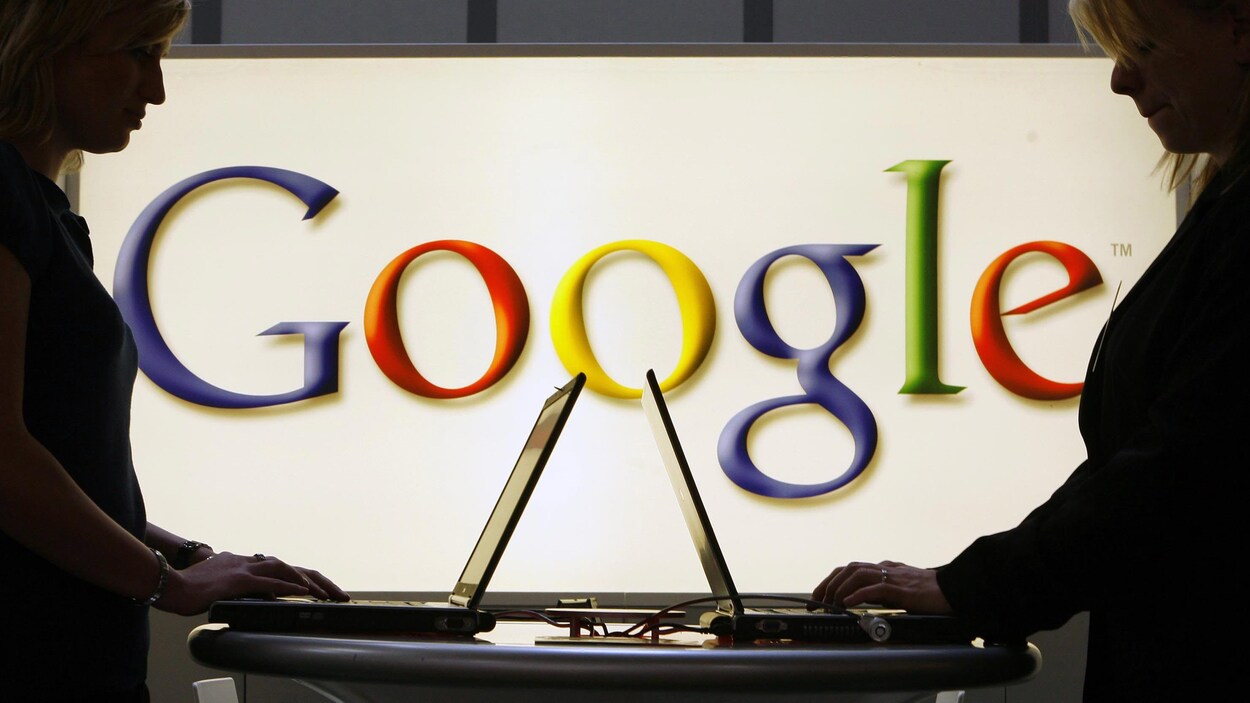 Deux femmes debout travaillent face à face sur des ordinateurs portables posés sur une table. En arrière-plan, le logo de Google est visible sur un écran lumineux.