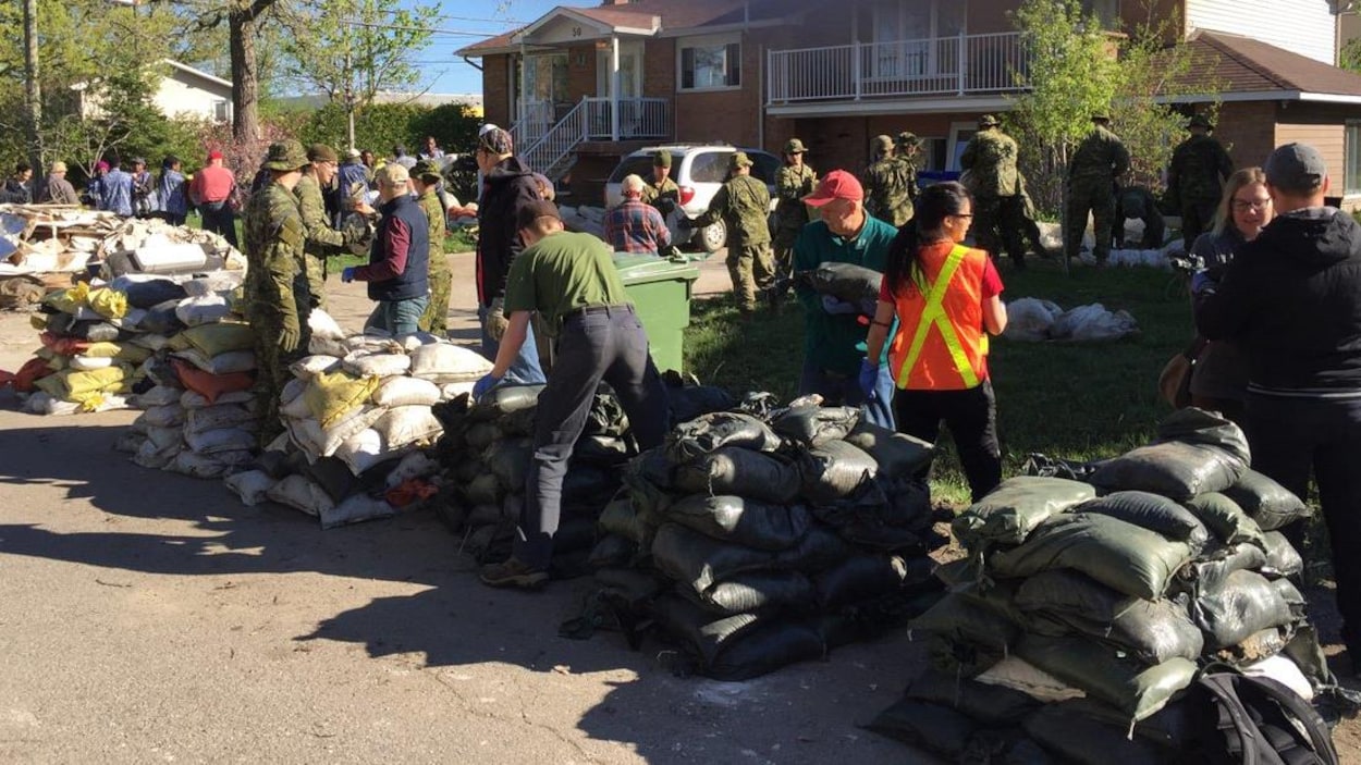Des militaires et des bénévoles ramassent des sacs de sable devant une maison.