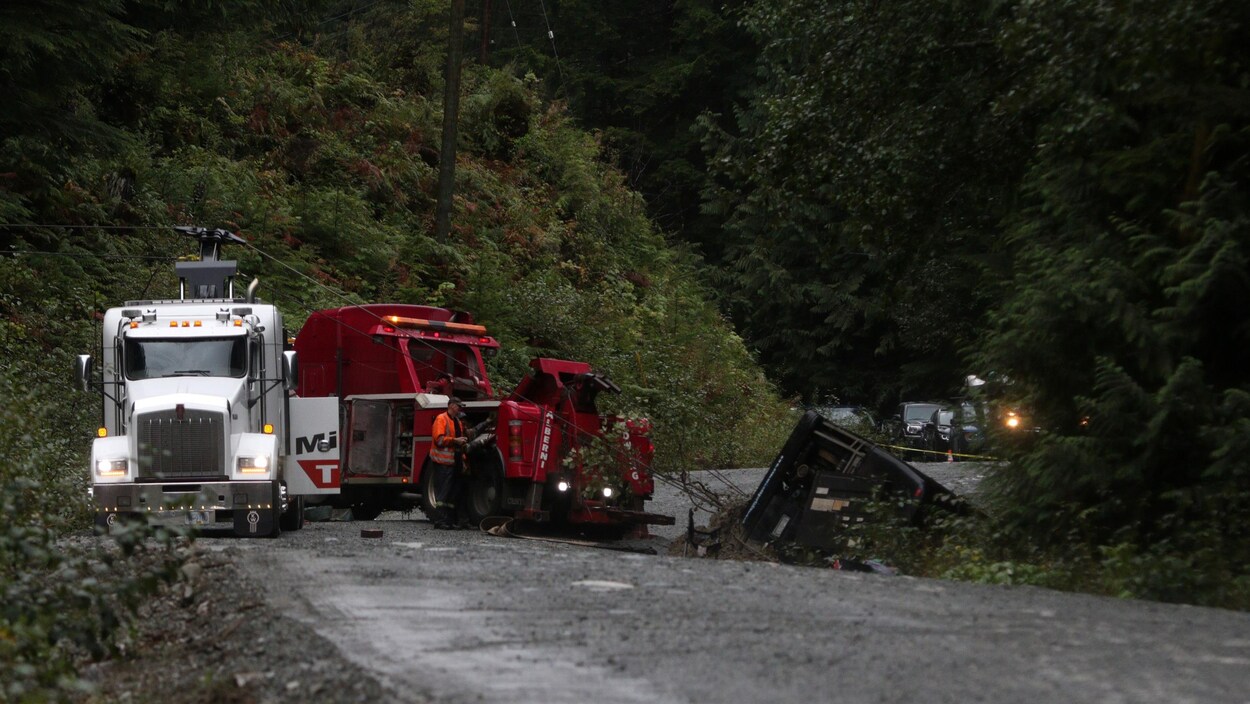 Une dépanneuse pour véhicules lourds sur une route de gravier à côté de laquelle se trouve un homme qui en opère le dispositif de remorquage pour actionner les câbles accrochés à l'autocar qui lui se trouve dans le fossé