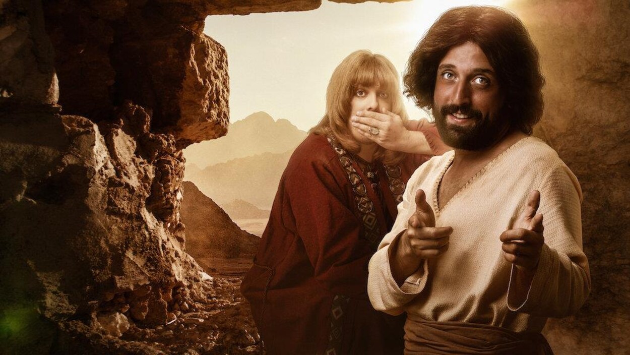 L'homme sourit en pointant devant lui. Une personne se cache la bouche derrière lui.