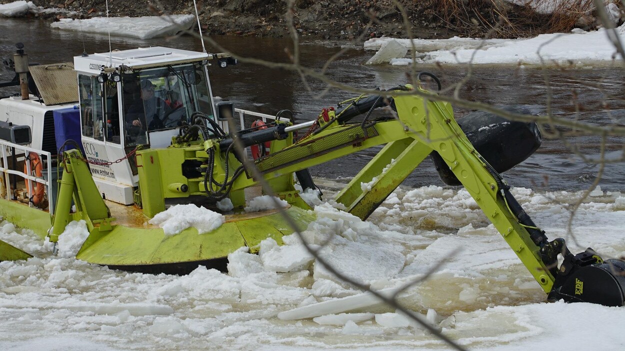 Une pelle mécanique flottante verte brise la glace dans la rivière.