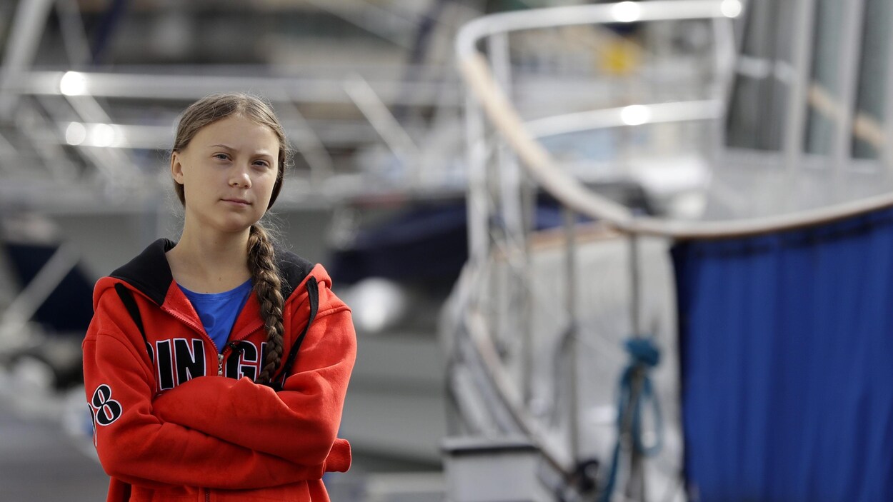 On voit l'adolescente, les bras croisés et le regard sérieux, qui pose devant des voiliers à quai.