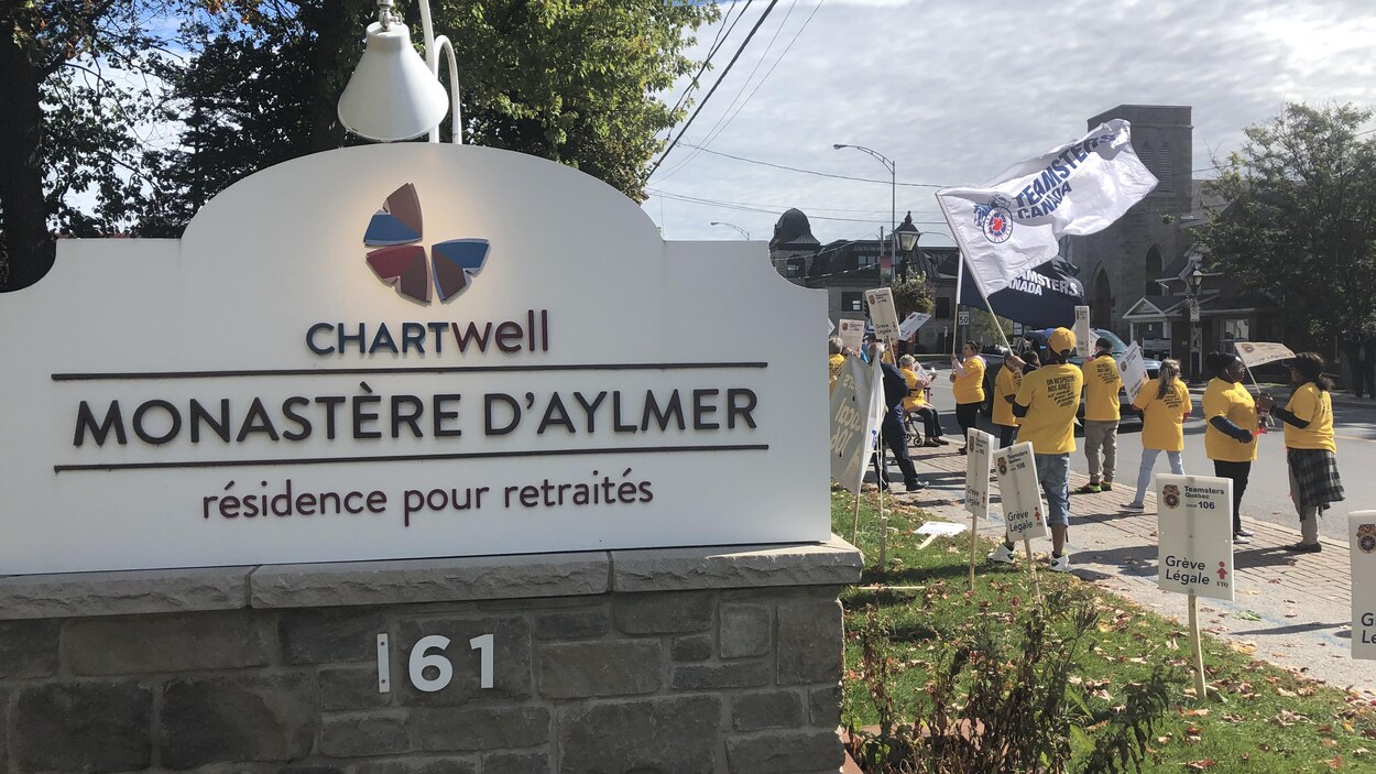 Des employés vêtus de chandails jaunes manifestent avec des drapeaux et des pancartes devant l'entrée de la résidence.