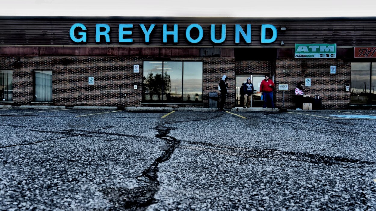 Entrée de la gare avec 4 personnes qui attendent devant sur le stationnement. Le logo Greyhound est affiché en haut.