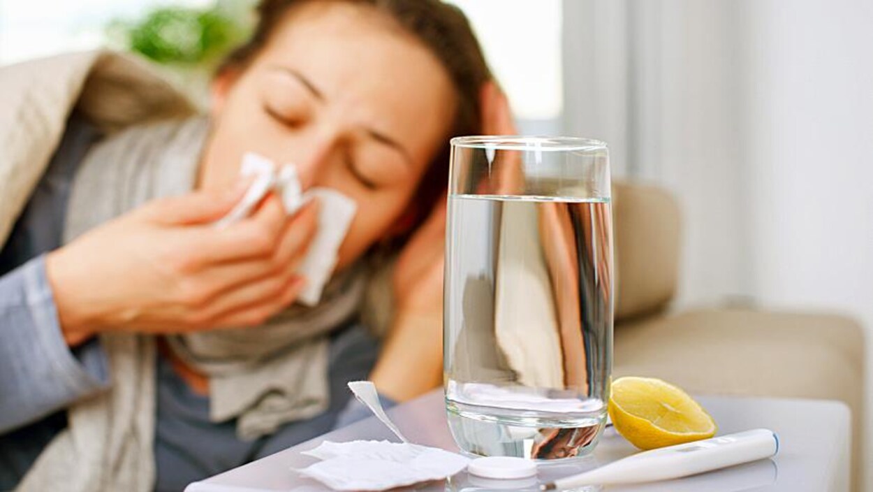 Une femme est couchée près d'un verre d'eau, d'un thermomètre et d'un morceau de citron. Elle semble éternuer dans un mouchoir et est visiblement mal en point.