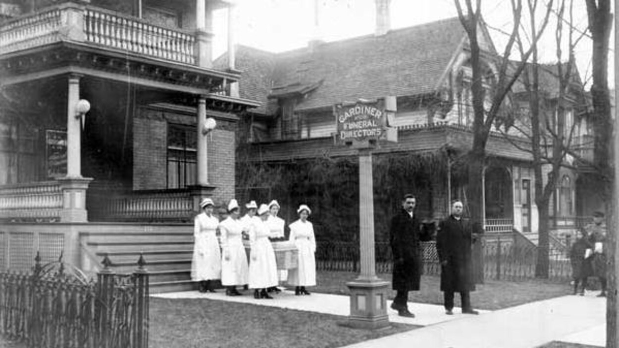 Sur une photo en noir et blanc, on voit six infirmières qui portent un cercueil, sortant d'un bâtiment et devancées par deux hommes habillés de longs manteaux noirs.