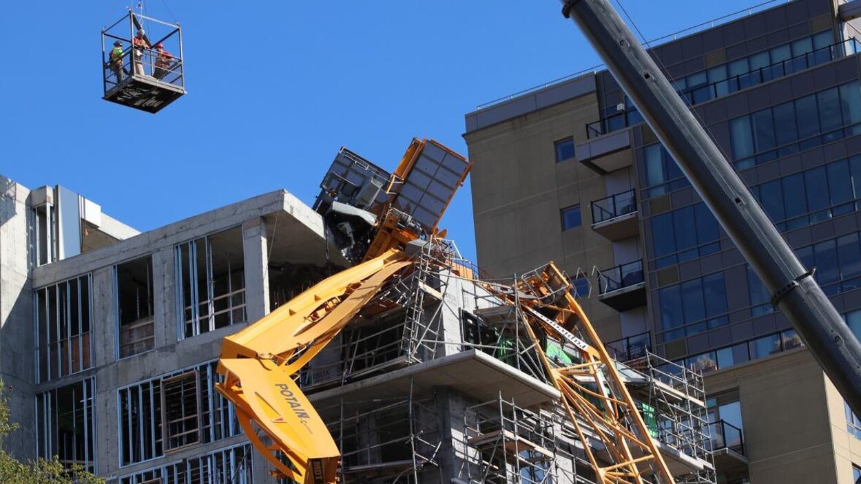 Trois ouvriers dans une nacelle passent au-dessus de la grue affaissée sur le toit de l'édifice.