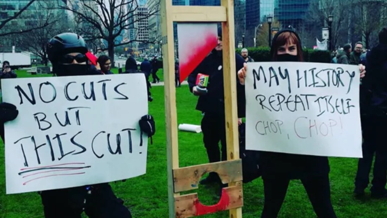 Des manifestants vêtus de noir et masqués tiennent des cartons où on peut lire des messages dénonçant les compressions.