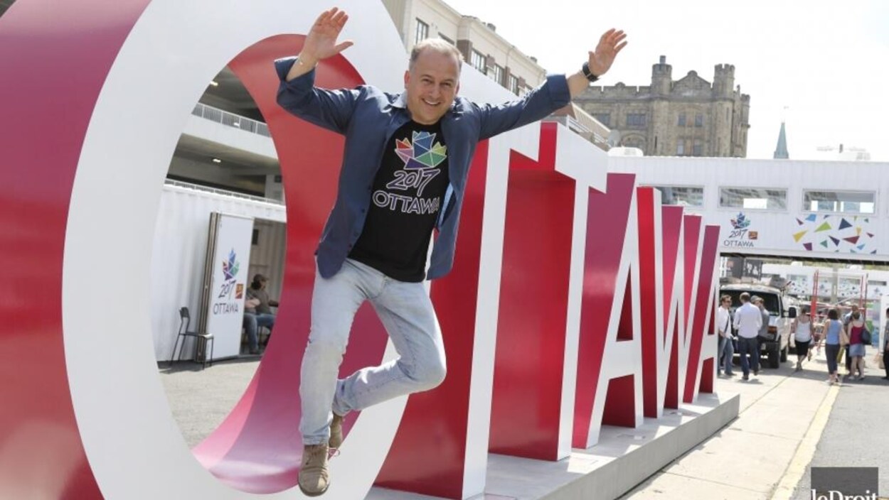 Guy Laflamme saute d'une lettre géante en forme de O, les bras dans les airs. Il est écrit Ottawa en lettres géantes. Guy Laflamme saute d'une lettre géante en forme de O, les bras dans les airs. Il est écrit Ottawa en lettres géantes.