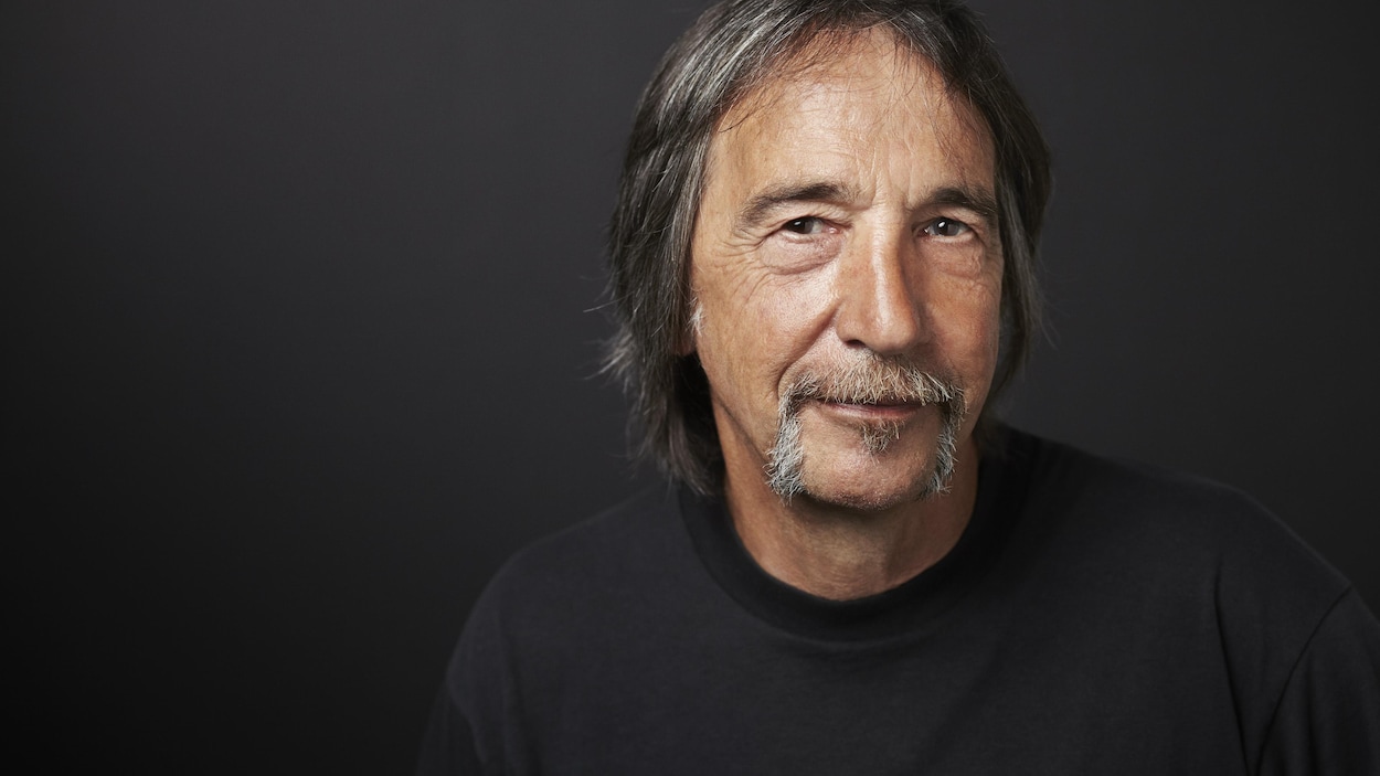 Portrait en couleur de Guy Lalancette sur fond gris foncé. Il regarde la caméra et esquisse un sourire. Il porte la moustache et les cheveux un peu longs.
