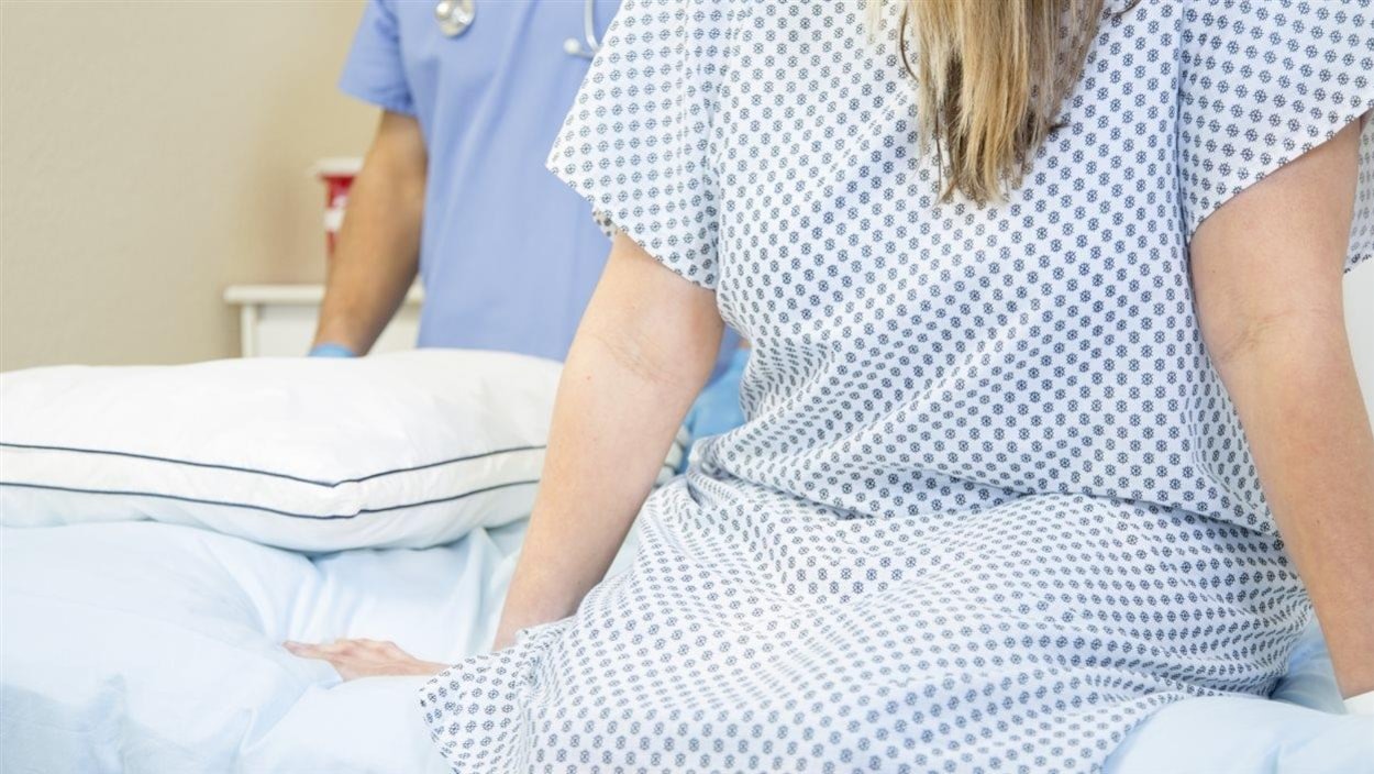 Une patiente en jaquette médicale assise sur un lit d'hôpital. On aperçoit un homme tenue médicale avec un stéthoscope derrière elle. Une patiente en jaquette médicale assise sur un lit d'hôpital. On aperçoit un homme tenue médicale avec un stéthoscope derrière elle.