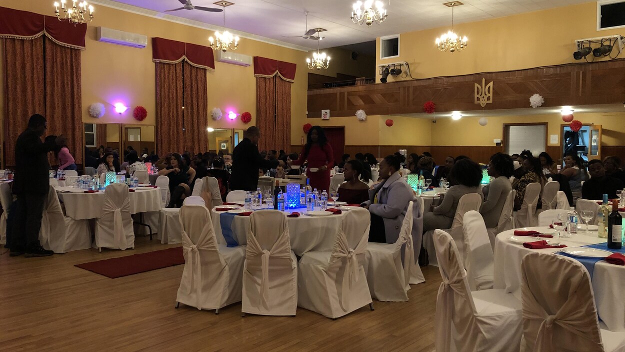 Des gens sont installés à des tables rondes dans une salle des fêtes.