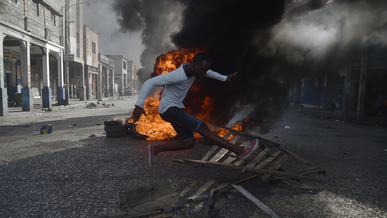 Une homme saute par dessus du bois calciné, un feu se consume au milieu d'une route derrière lui.