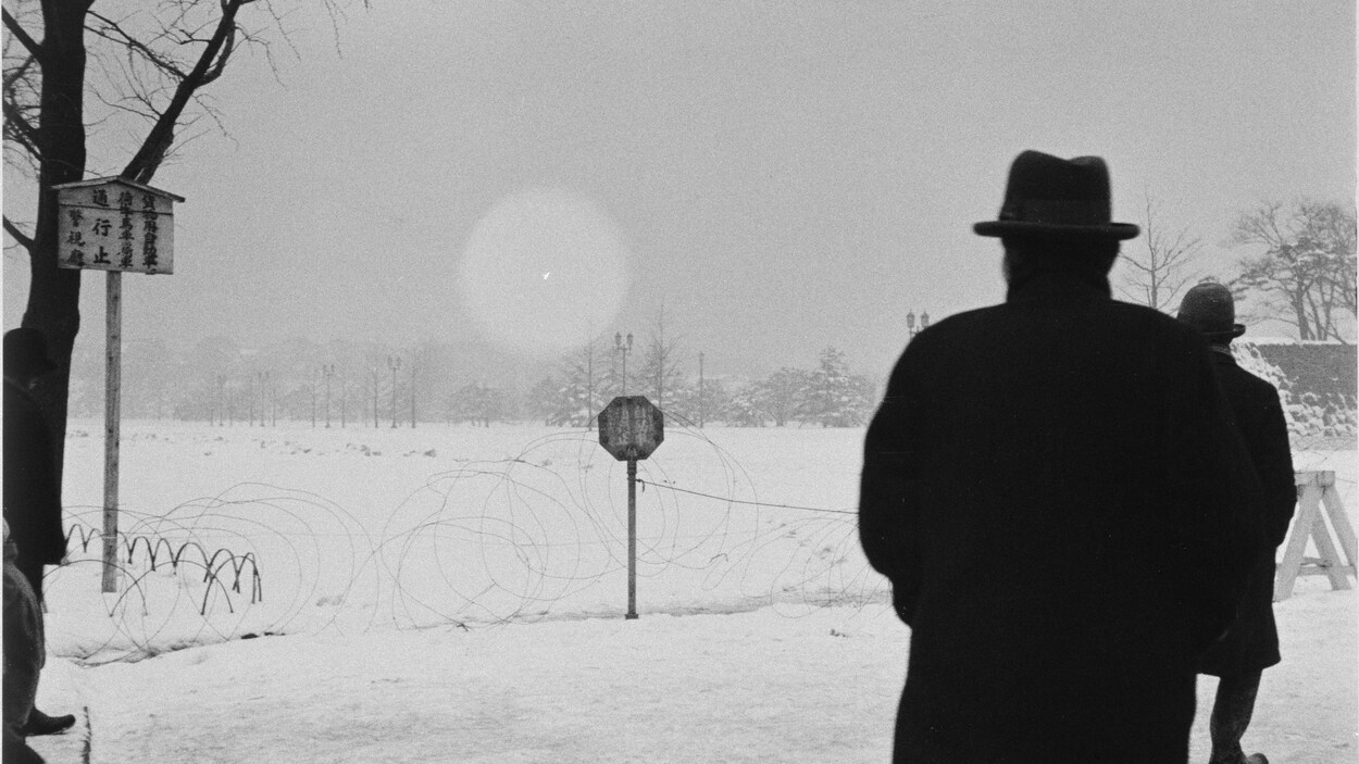 Une photographie en noir et blanc montre la silhouette d'un homme vêtu d'un grand manteau et d'un chapeau devant un arbre.