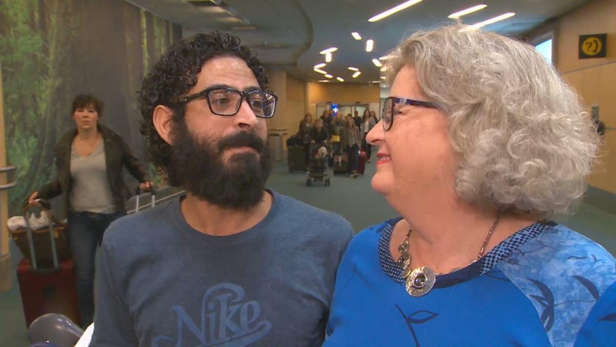 Un homme avec des cheveux frisés noirs, une moustache et une barbe touffue et des lunettes à monture noire regarde avec amour une femme âgée blonde qui le regarde avec un sourire béat.