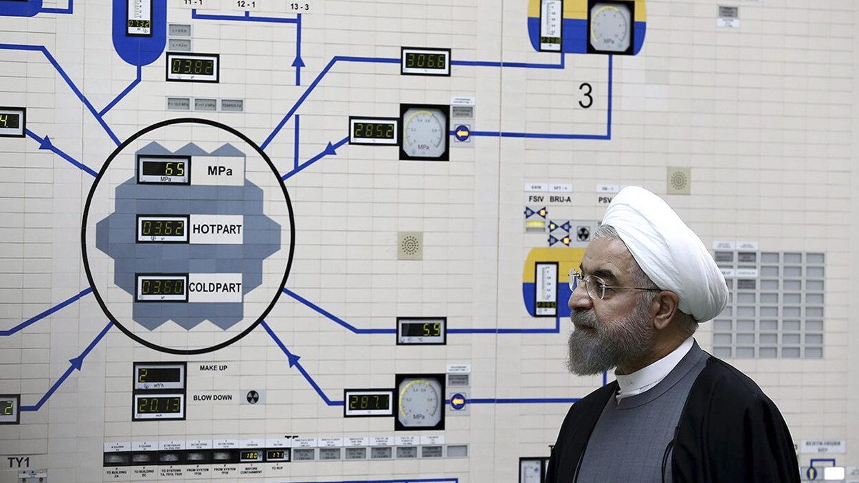 Le président iranien, portant la barbe mi-longue, des lunettes et un chapeau enturbanné, observe un mur où figure un diagramme et des cadrans analogiques.