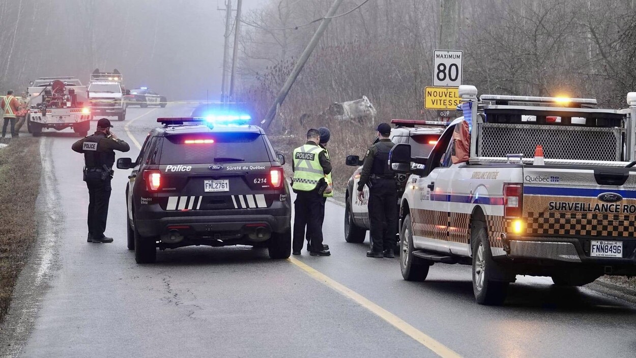 Des voitures de la Sûreté du Québec et une remorqueuse sur une route. À droite on voit un poteau brisé et un véhicule accidenté, à l'envers.