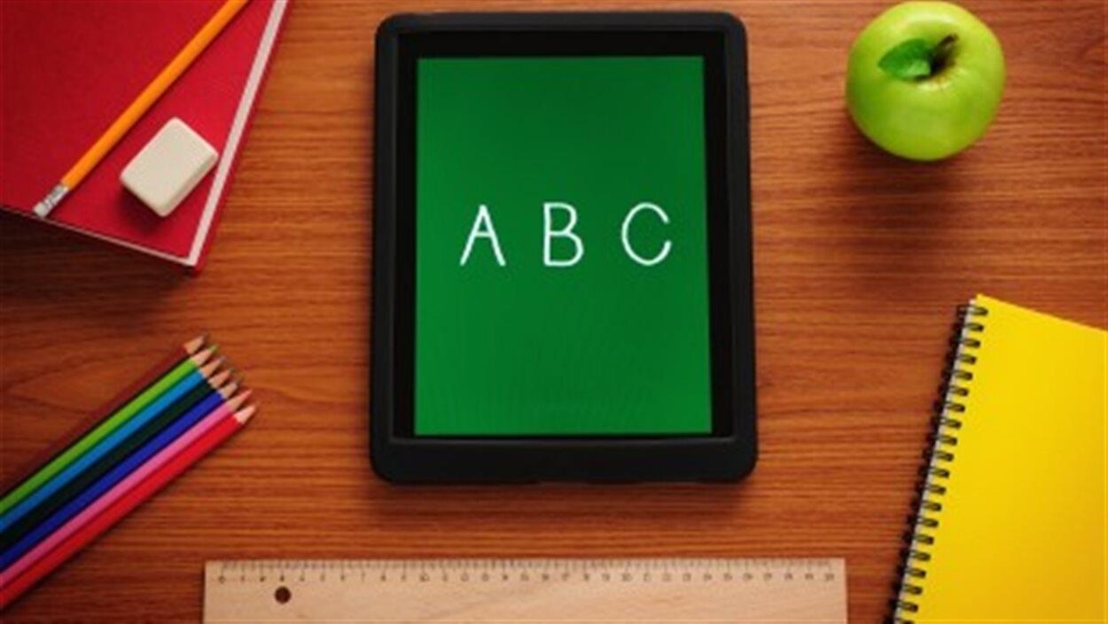 Une tablette électronique affichant les lettres A, B et C posée sur un pupitre d'école en bois et entourée de fournitures scolaires.